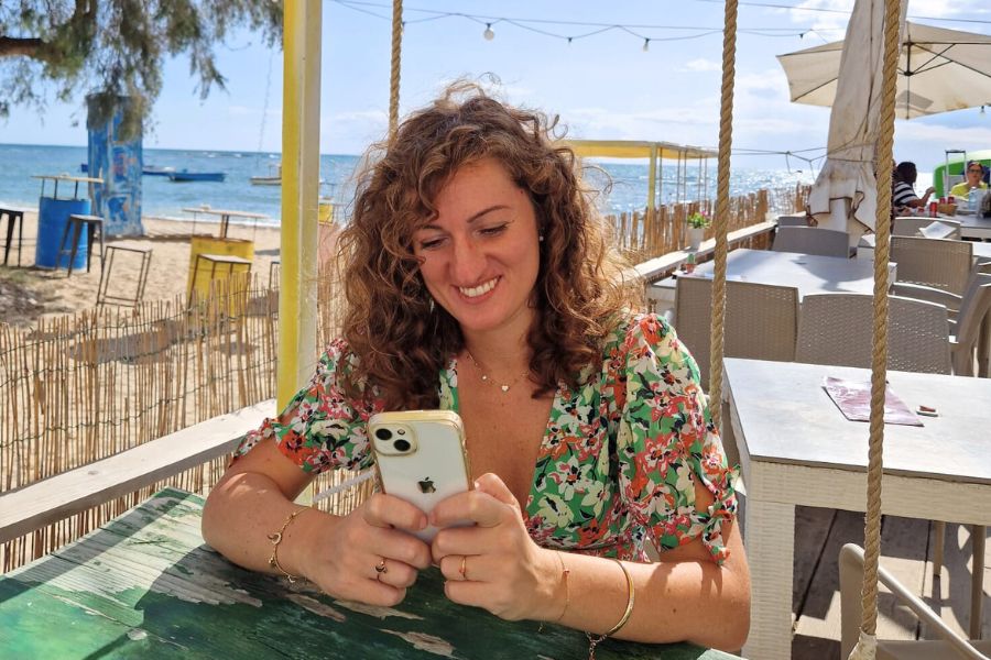 Foto di Maria Grazia De Falco seduta a un tavolo con il mare di sfondo, intenta a guardare lo smartphone mente sorride