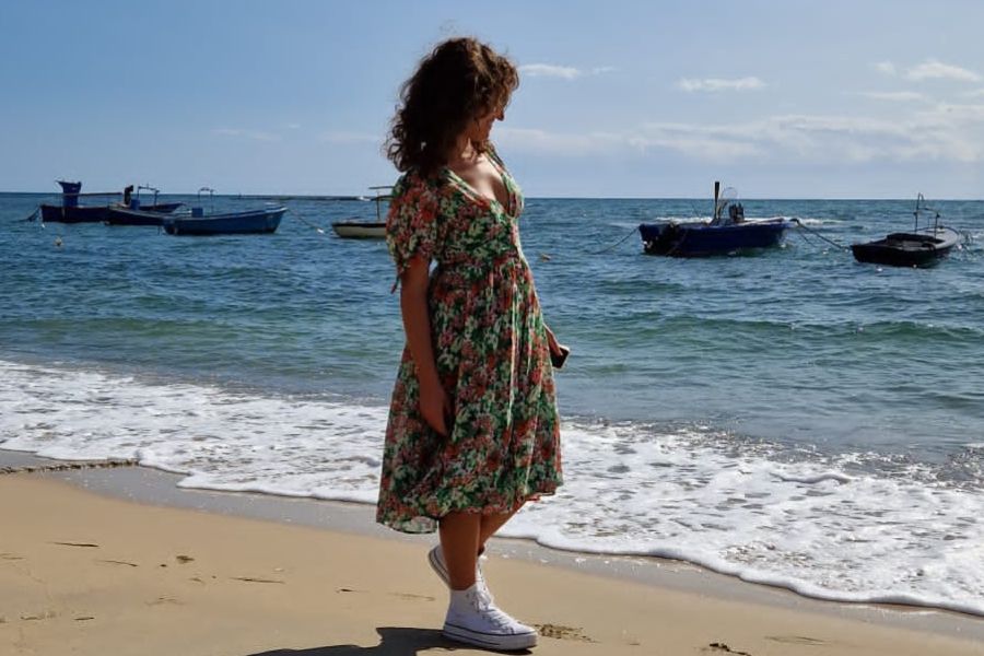 Ragazza con vestito verde a fiori che cammina sulla riva del mare. sullo sfondo, delle barche arenate in acqua