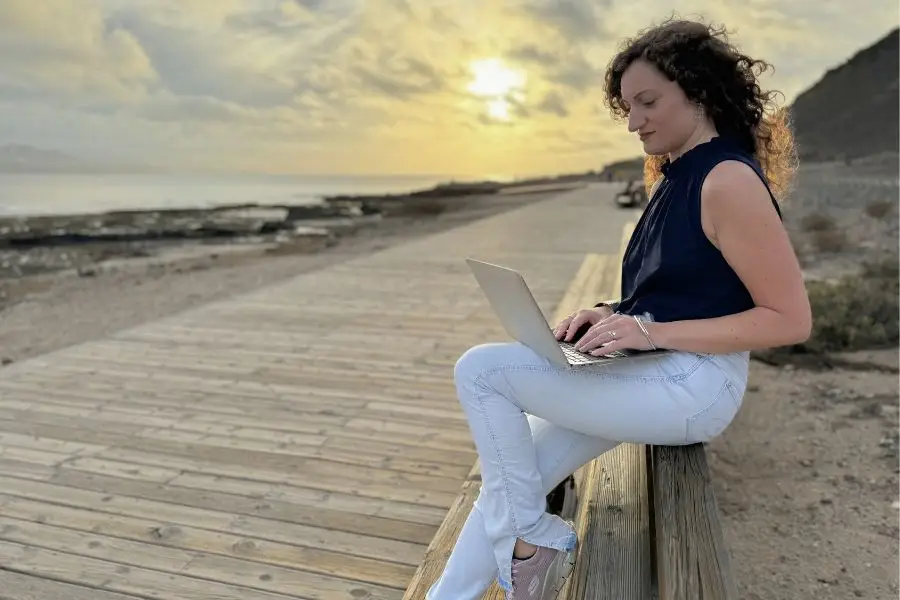 ragazza seduta su uno schienale di una panchina, con il computer poggiato sulle gambe. il tramonto sullo sfondo